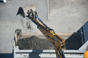 An excavator boom arm with a bucket dropping ground material into a steel bin